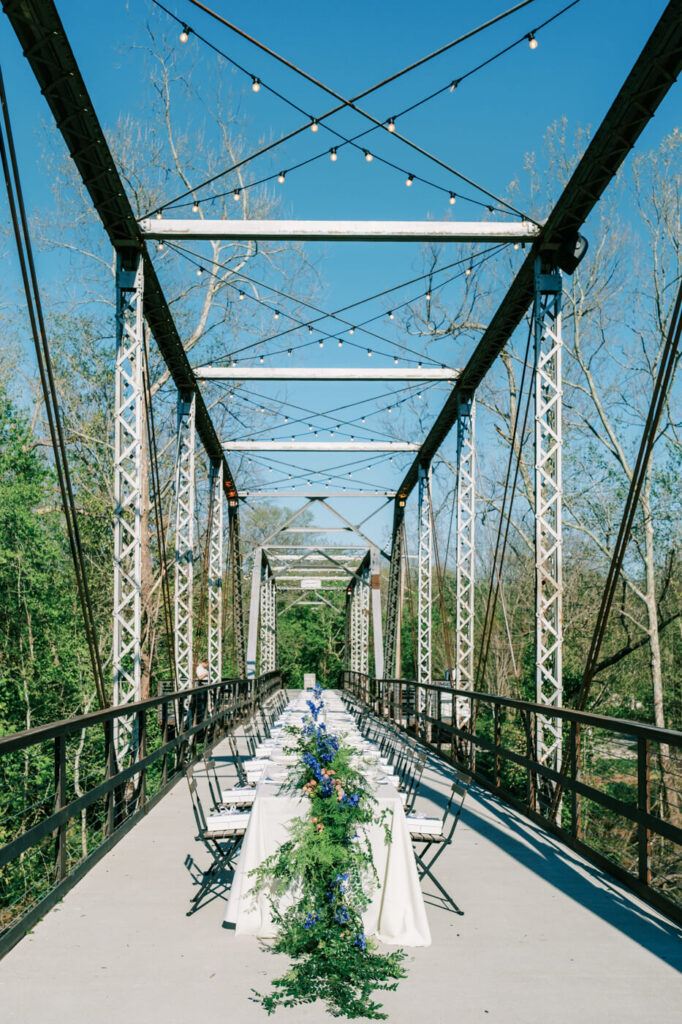 Rehearsal dinner table on Finley Farms Bridge – A stunning long dining table adorned with blue and green floral arrangements set under the open sky on the historic bridge.