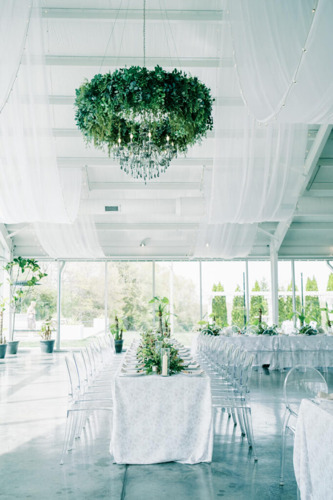 Reception at The Greenhouse Two Rivers with greenery-wrapped chandelier and draped ceiling – A bright reception space at The Greenhouse Two Rivers, featuring a stunning chandelier wrapped in fresh greenery, flowing white fabric draped across the ceiling, and elegantly set long tables.