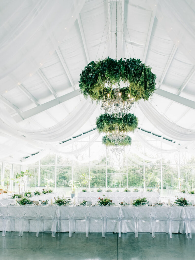 Whimsical reception decor with greenery chandeliers at Two Rivers Greenhouse – Two cascading greenery chandeliers hang above long reception tables, draped in white linens and surrounded by clear ghost chairs at Two Rivers Greenhouse.