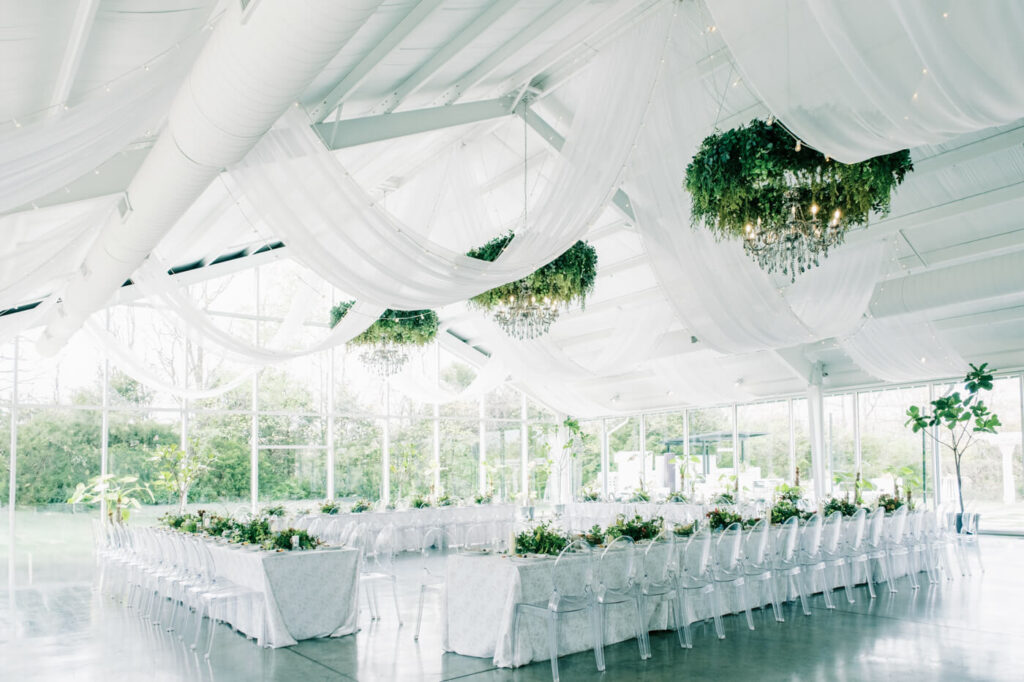 Light-filled reception at Two Rivers Greenhouse wedding venue – A breathtaking reception setup featuring elegant draping, lush greenery chandeliers, and natural light streaming through the glass walls of Two Rivers Greenhouse.