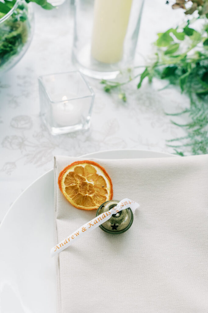 Personalized wedding place setting at Two Rivers Greenhouse – A custom name tag tied to a delicate bell, accompanied by dried citrus and soft candlelight, creating a meaningful and intimate guest experience at a Two Rivers Greenhouse wedding.