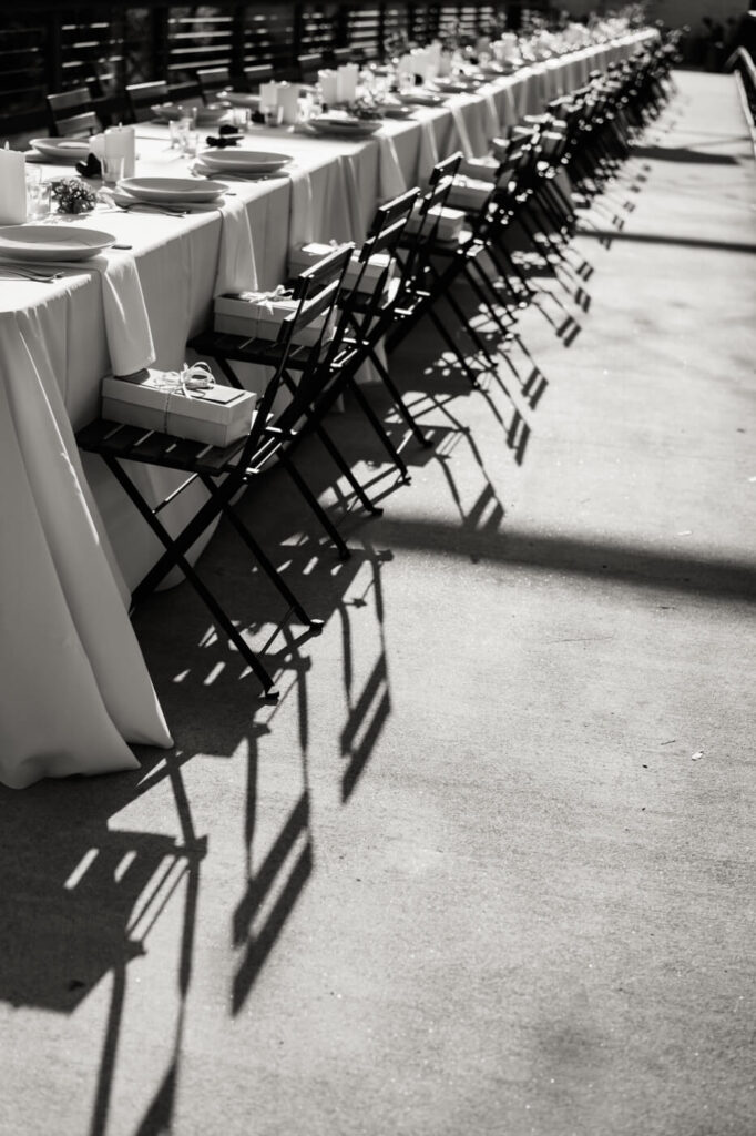 Black and white image of the rehearsal dinner setup – The golden-hour light casts dramatic shadows on the row of neatly arranged chairs and place settings at Finley Farms Bridge.