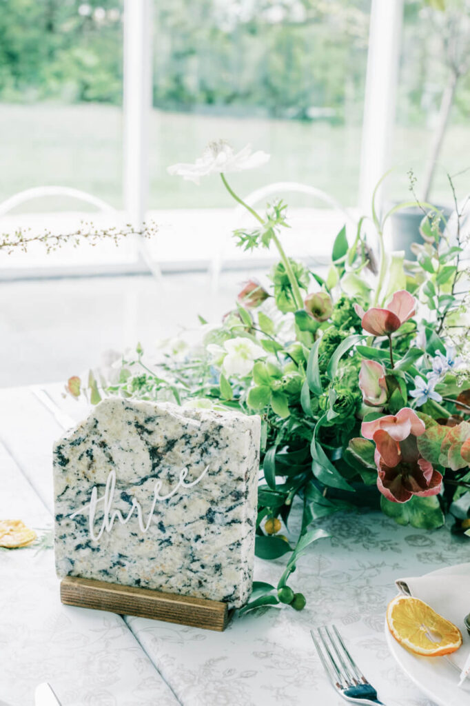 Granite table number at Greenhouse Two Rivers wedding – A rustic granite table number with delicate white script reading "three," placed beside lush floral arrangements at Greenhouse Two Rivers in Missouri.