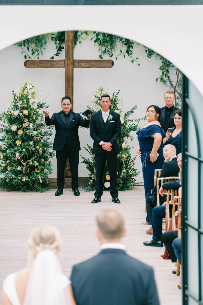 Groom awaits the bride at Greenhouse Two Rivers ceremony – The groom stands at the altar beneath a wooden cross, surrounded by lush floral arrangements, as he watches his bride approach at Greenhouse Two Rivers.