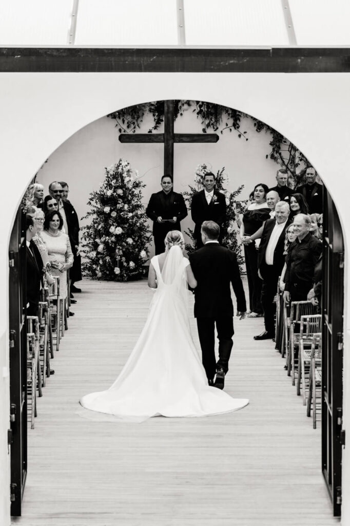 Bride walks down the aisle at Greenhouse Two Rivers wedding – A black and white image of the bride and her father walking down the aisle toward the groom at Greenhouse Two Rivers.