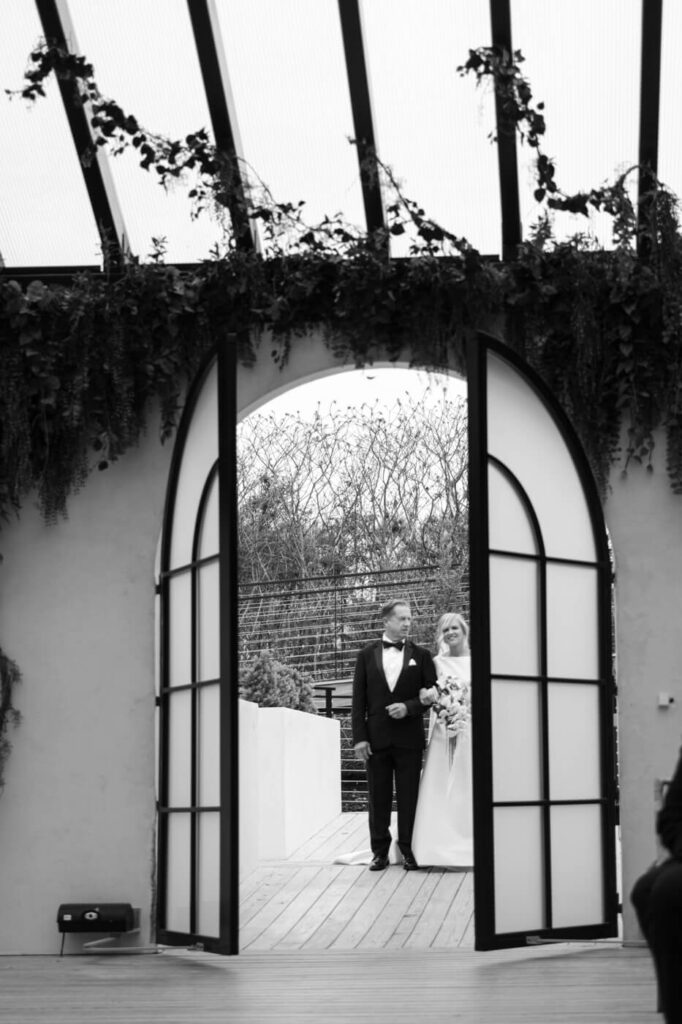 Bride and father entering Greenhouse Two Rivers ceremony space – A timeless black and white shot of the bride and her father stepping through arched doors into the wedding ceremony at Greenhouse Two Rivers.