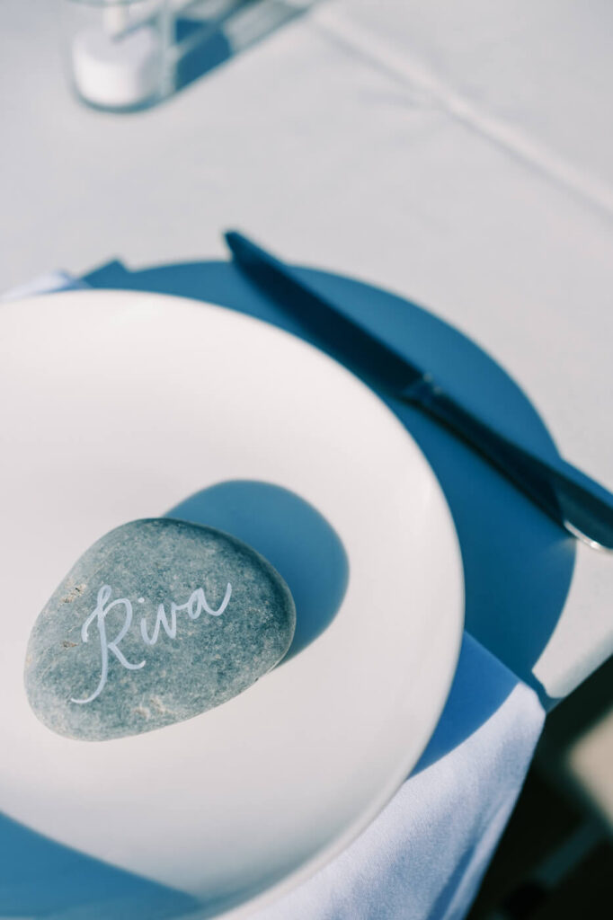 Personalized stone place card at Finley Farms Bridge – A hand-lettered stone place card reading "Riva" sits atop a white plate, adding a unique touch to the rehearsal dinner.