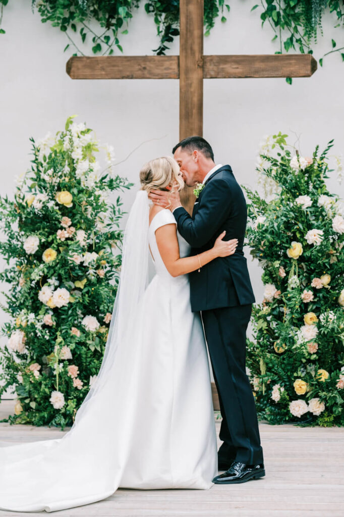 Bride and groom sharing a heartfelt kiss in front of a wooden cross, framed by lush floral arrangements.