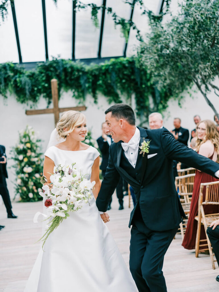 Bride and groom celebrating as they walk down the aisle, the groom playfully leaning toward his bride with joy.