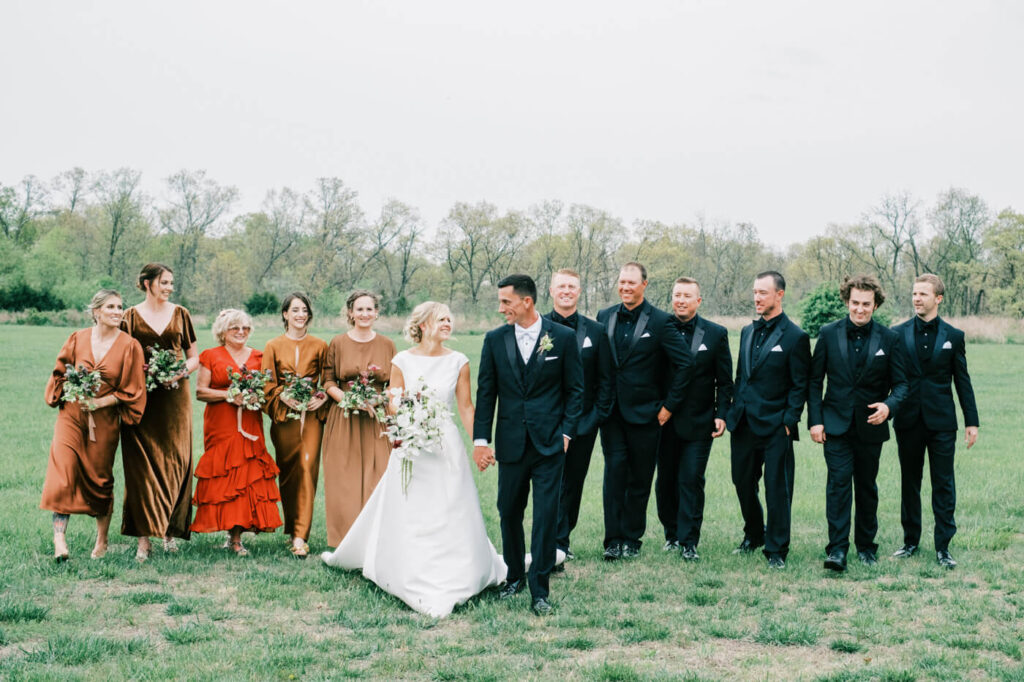 Wedding party walking together in an open field, bridesmaids in earthy-toned dresses and groomsmen in black suits.