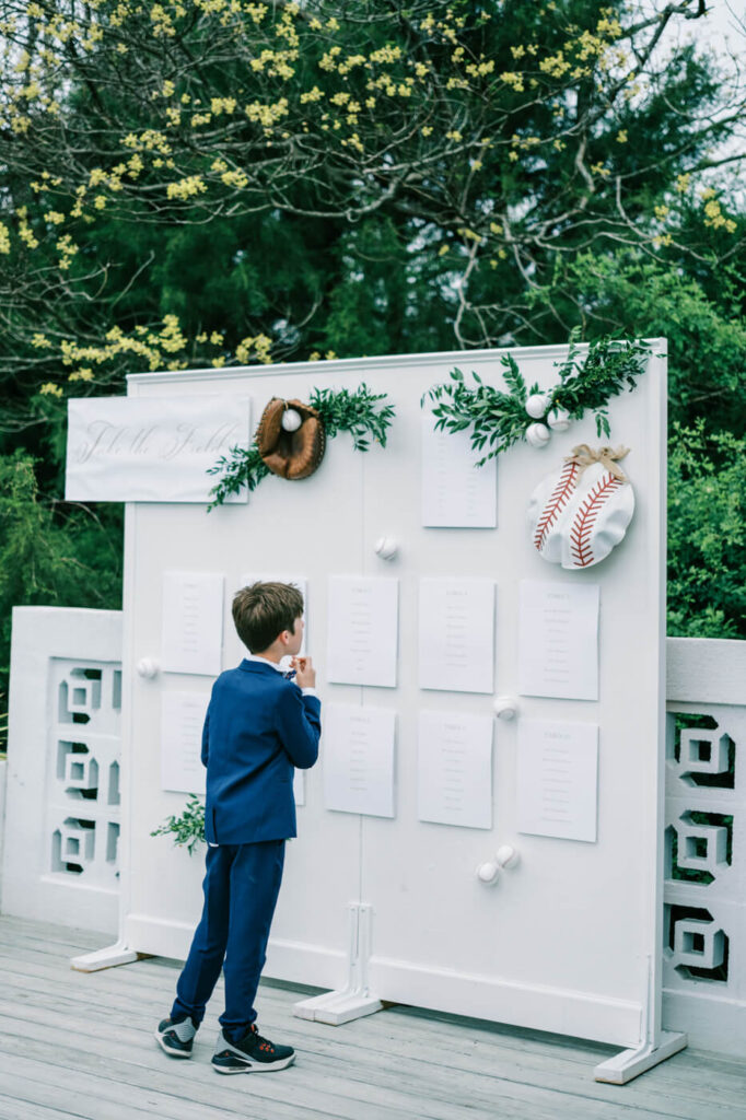 A young boy in a navy suit examines a seating chart decorated with baseball-themed elements and greenery.