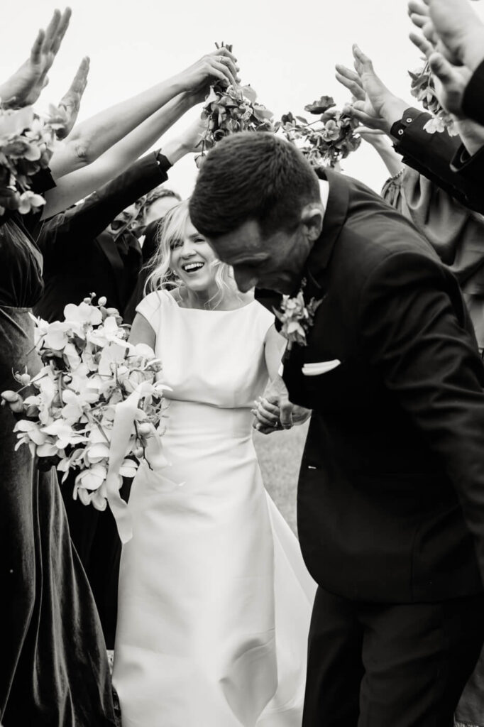 Black and white candid moment of the bride and groom laughing as they run under a floral arch formed by the wedding party.