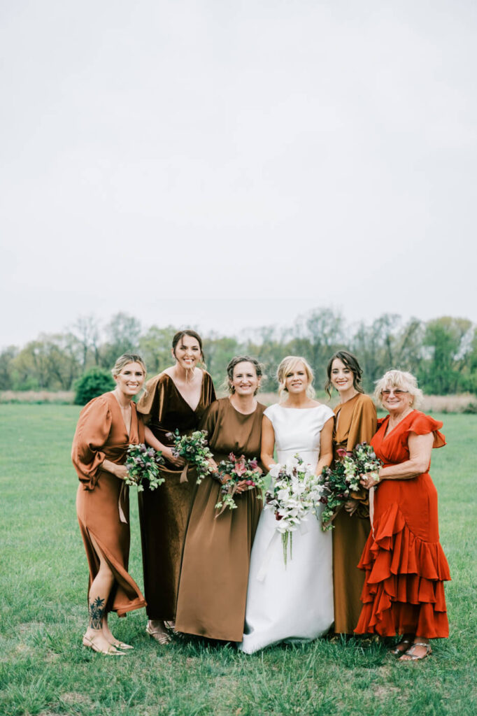 Bride posing with her bridesmaids in an open field, each holding a lush floral bouquet in warm tones.