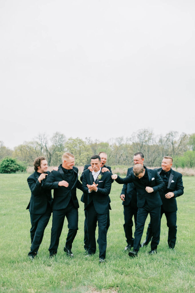 Groom in black suit being playfully teased by his groomsmen, all laughing in an open grassy field.