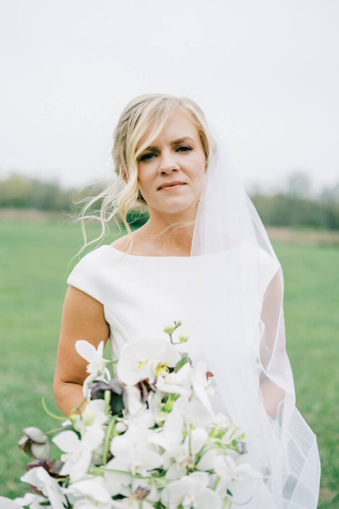 Bride gazing softly into the camera, her sheer veil draped over her shoulder as she holds a bouquet of white orchids.