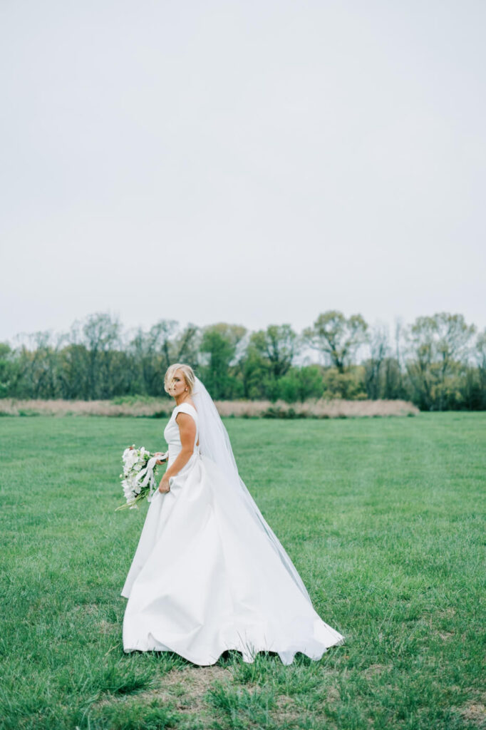 Bride in Flowing White Gown - The bride gracefully walks across a vast green field, her elegant white dress billowing behind her as she holds her cascading bouquet.