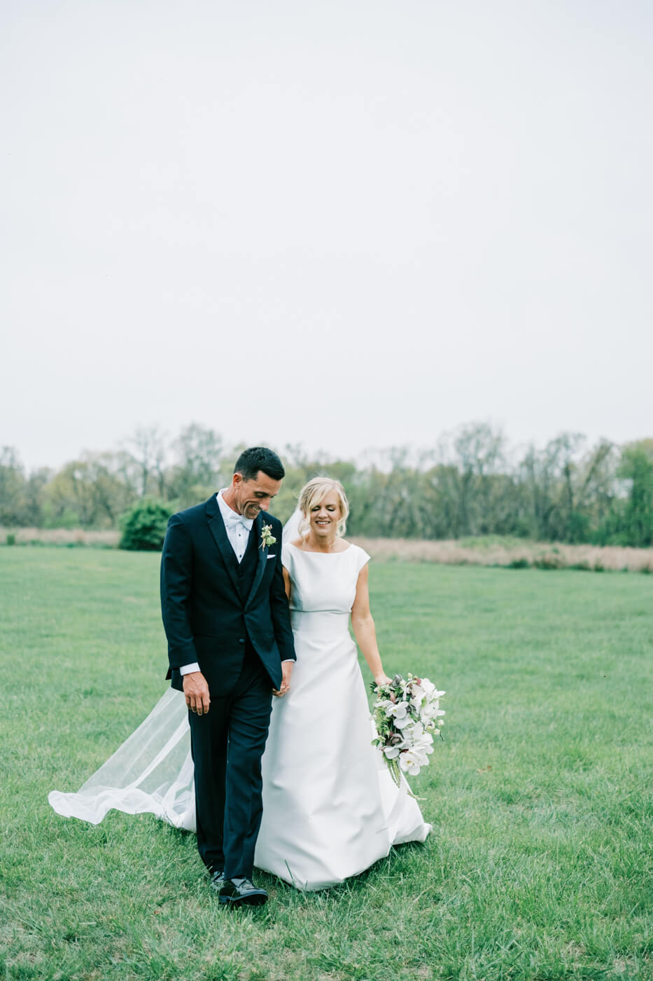 Bride and Groom Walking in a Field at Greenhouse Two Rivers – A joyful moment as the newlyweds walk hand in hand through a lush green field at Greenhouse Two Rivers, the bride's elegant gown flowing behind her.