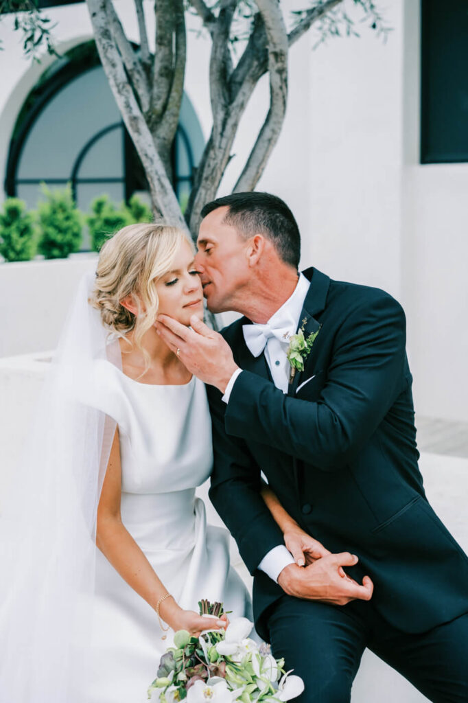 Romantic Whisper Under an Olive Tree at Greenhouse Two Rivers – The groom tenderly whispers to his bride as he gently cups her chin, sitting together beneath an olive tree at Greenhouse Two Rivers, surrounded by timeless beauty.