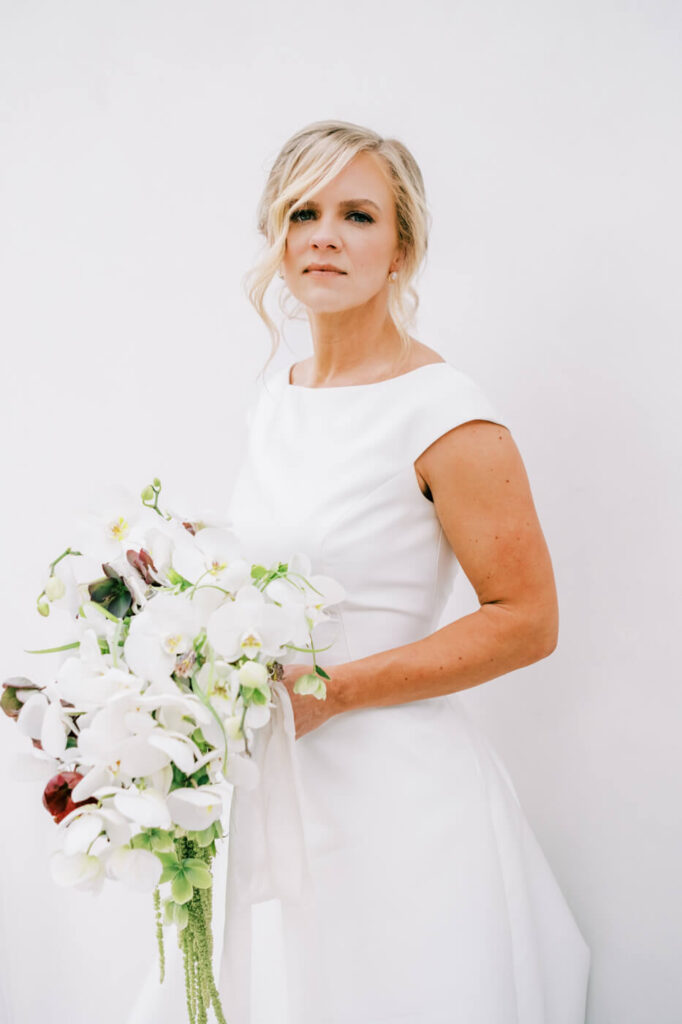 Classic Bridal Portrait at Greenhouse Two Rivers – The bride stands gracefully against a white backdrop at Greenhouse Two Rivers, holding a cascading bouquet of white orchids and burgundy blooms.