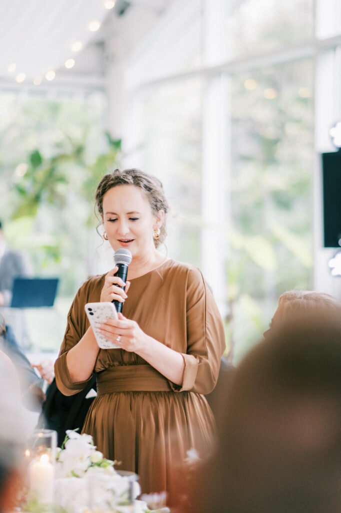 Maid of Honor Toast at Greenhouse Two Rivers – The maid of honor, dressed in a sophisticated brown gown, delivers a heartfelt speech while holding a microphone at Greenhouse Two Rivers.