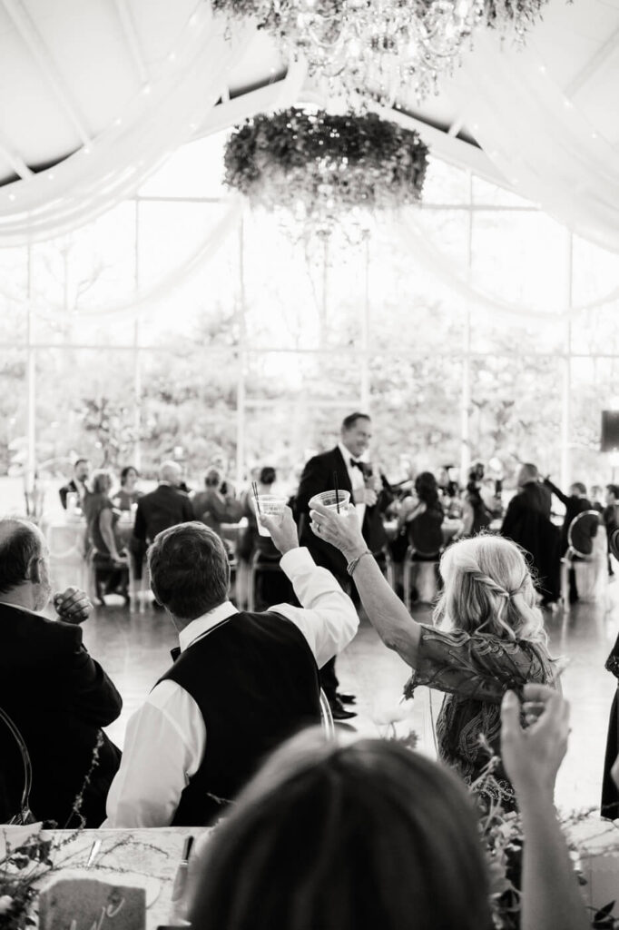 Cheers and Toasts at Greenhouse Two Rivers Wedding Reception – A black-and-white image of wedding guests raising their glasses in a celebratory toast inside the beautifully decorated Greenhouse Two Rivers venue.