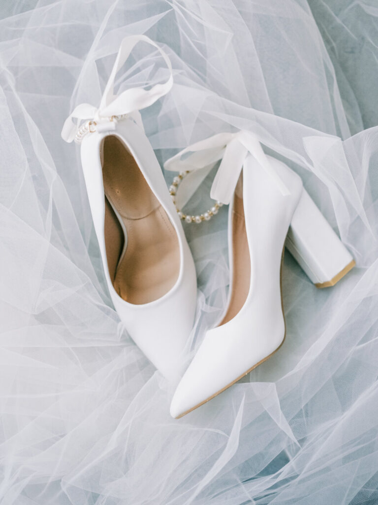 Bride’s White Wedding Shoes on Soft Tulle – A pair of elegant white wedding heels with delicate ribbon ties and pearl details, resting on soft layers of tulle, perfectly capturing the bride’s timeless style.