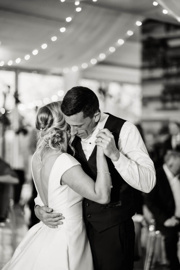 Bride and Groom First Dance Close-Up - A black-and-white image of the couple sharing their first dance, the groom lovingly holding the bride close under twinkling string lights.