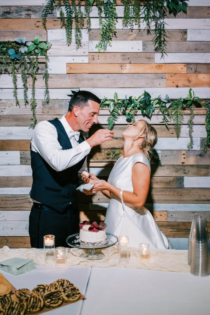 Cake Cutting Celebration - The bride and groom laugh as they feed each other cake, standing in front of a beautifully decorated rustic wooden wall adorned with greenery.