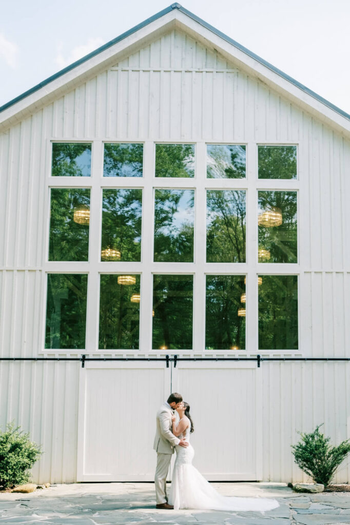 Bride and groom share a kiss in front of the white barn doors at Carolina Grove, with large windows reflecting the lush greenery of the venue.