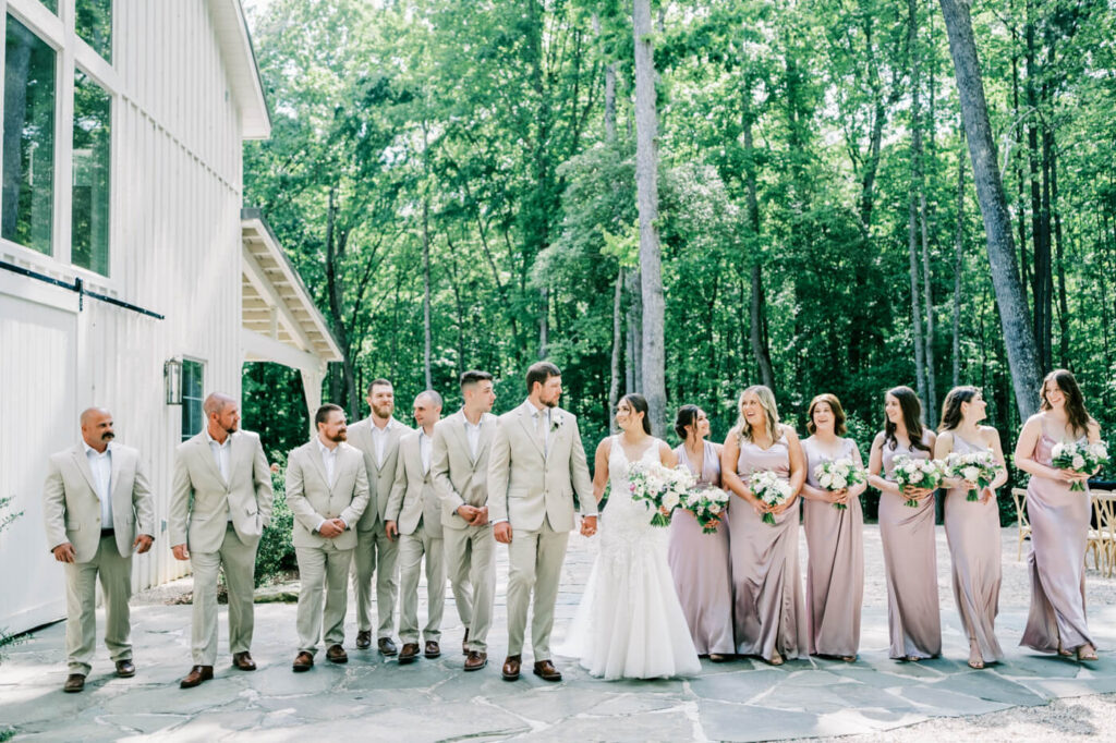 Wedding party walking together in front of the white barn at Carolina Grove, with bridesmaids in blush satin dresses and groomsmen in tan suits.