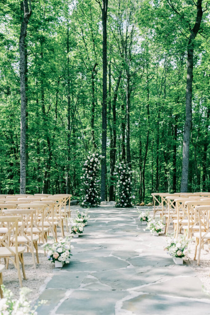 Outdoor wedding ceremony setup at Carolina Grove, featuring wooden chairs, a stone aisle, and a floral arch surrounded by lush forest.