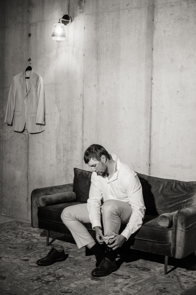 Groom Aiden getting ready in a modern rustic suite, adjusting his shoes in a timeless black-and-white moment.