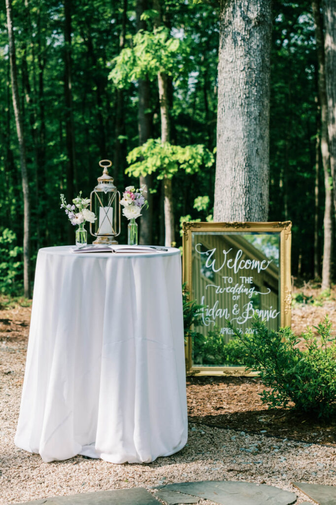 Wedding welcome table setup with a white linen table, floral arrangements, and a gold-framed welcome sign in the wooded venue at Carolina Grove.