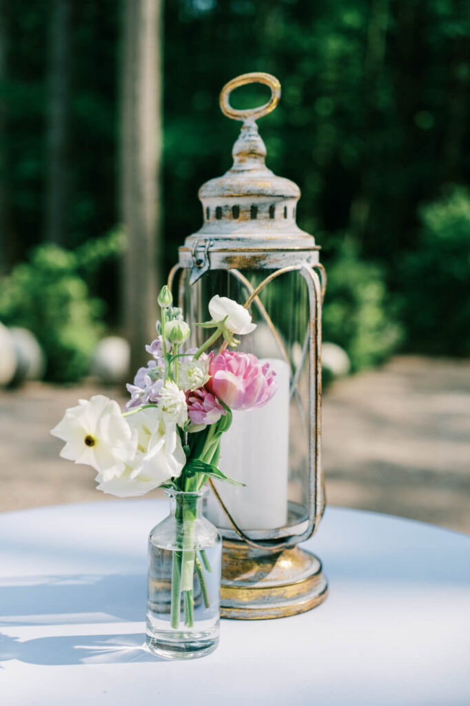 Close-up of a vintage lantern and delicate floral arrangement in a glass vase on a white table, exuding a romantic and timeless wedding aesthetic.