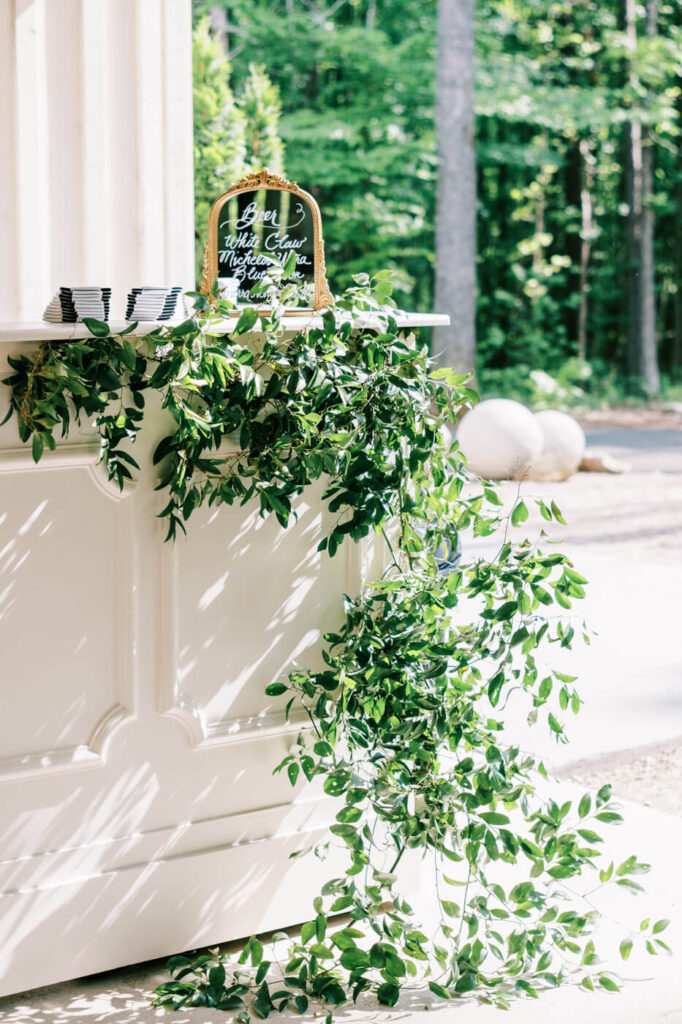 Lush greenery cascading over a white bar with a gold-framed drink menu sign, bathed in dappled sunlight at an outdoor wedding reception.