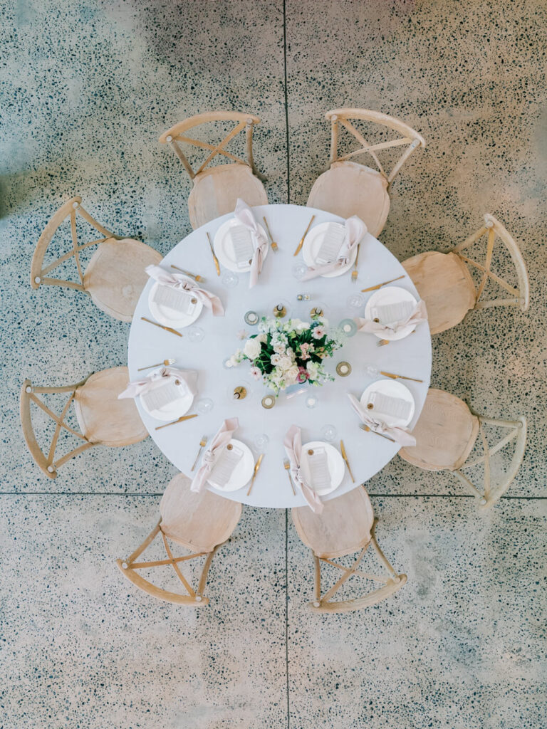 A beautifully styled reception table with natural wood chairs, white linens, and delicate floral centerpieces captured from an overhead perspective.