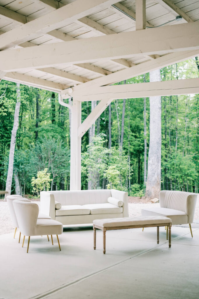 Elegant white lounge seating under a covered pavilion, offering a cozy and refined wedding reception space with lush greenery in the background.