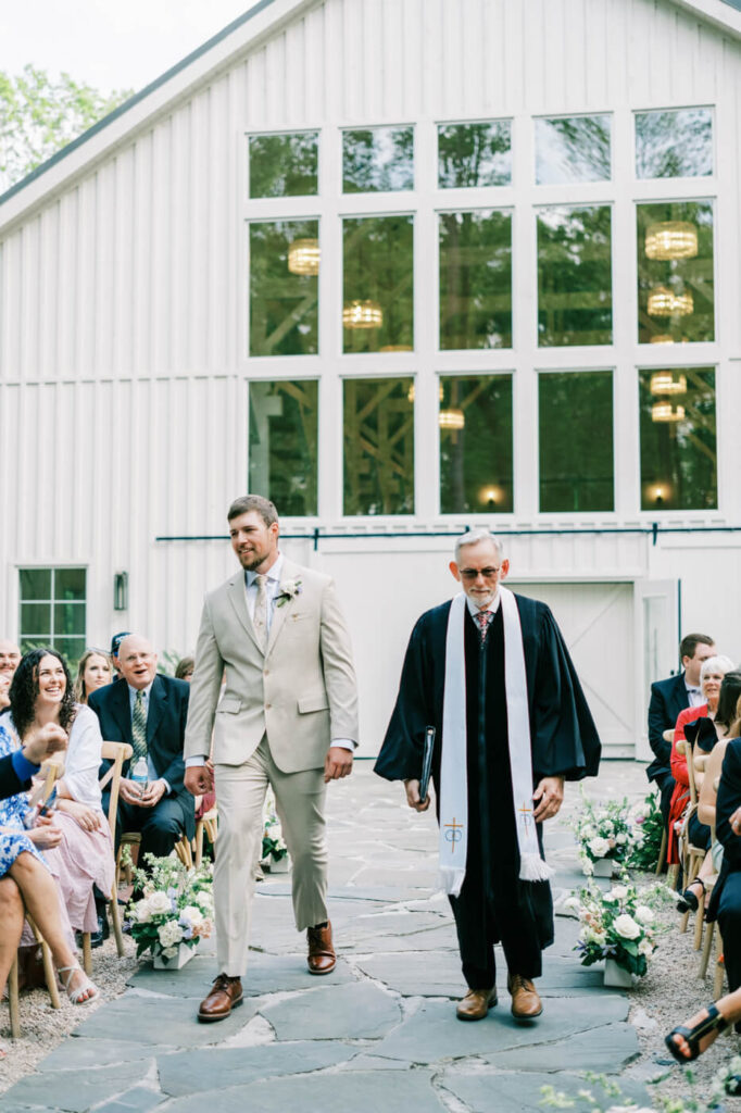 The groom, dressed in a light beige suit, walking down the stone aisle with the officiant in front of the white barn venue at Carolina Grove.