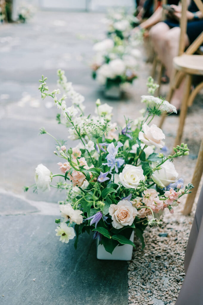 Aisle Floral Arrangements – Lush floral arrangements of blush, white, and lilac blooms line the ceremony aisle on stone pathways.