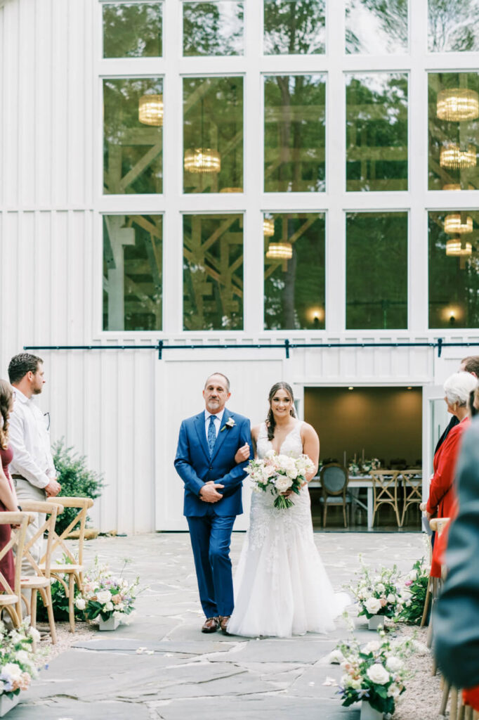 Bride Walking Down the Aisle – A beaming bride in a lace wedding gown walks arm-in-arm with her father, surrounded by guests and pastel floral arrangements.