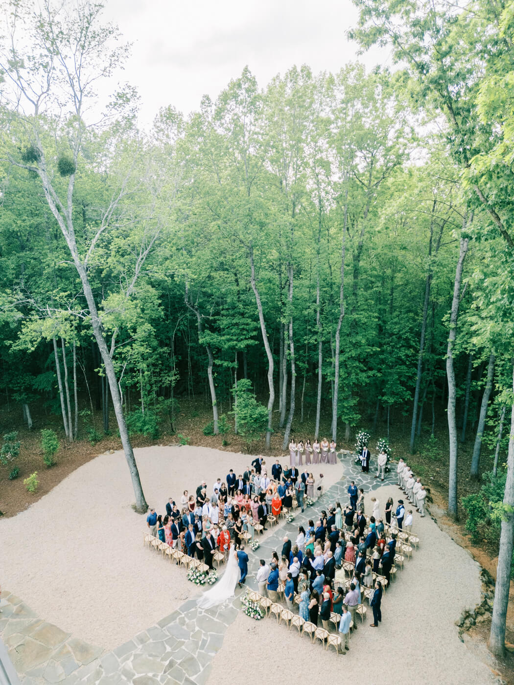 A breathtaking aerial view of Bonnie & Aiden's outdoor wedding ceremony at Carolina Grove, surrounded by lush greenery. Guests are seated in a U-shape, as the bride walks down the aisle toward the groom. The elegant barn venue and natural woodland setting create a romantic and timeless atmosphere.