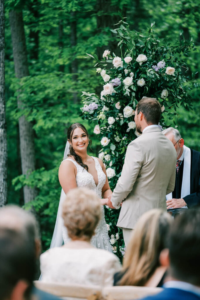 Bride Smiling at Groom – The bride, glowing with happiness, gazes at her groom during the vows, framed by a lush floral arch.