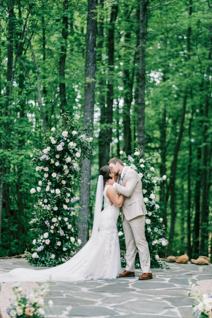 The First Kiss – The newlyweds share their first kiss under a floral arch, framed by the natural greenery of the forest.