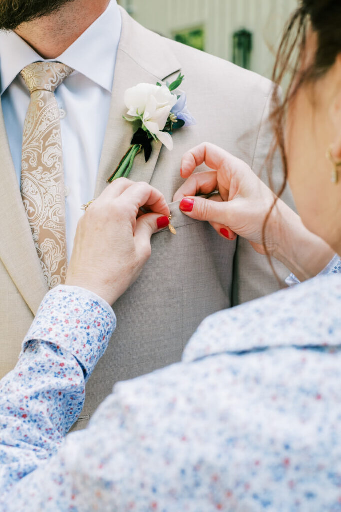 Mother of the groom pinning a classic boutonnière on Aiden’s beige suit, a heartfelt moment before the ceremony.