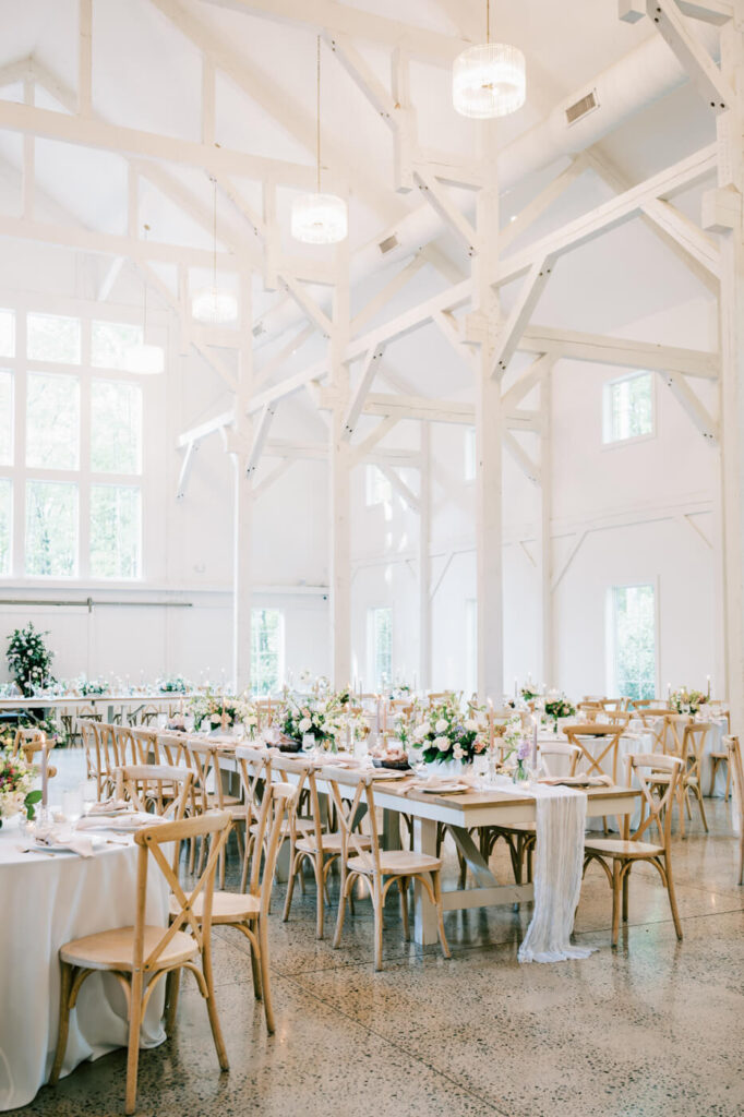 A beautifully set reception table with wooden cross-back chairs, blush-toned napkins, delicate floral arrangements, and glowing candlelight in an airy white barn with large windows.