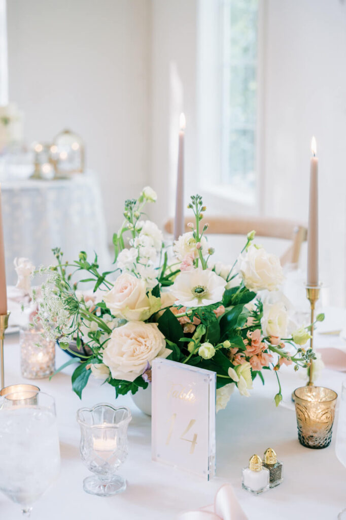 A close-up of a floral centerpiece featuring pastel roses, greenery, and white blooms, surrounded by soft candlelight and glassware on a reception table.