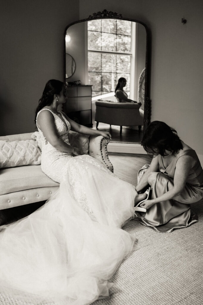 Bride Bonnie sits elegantly as her mother helps her with shoes, reflected in a grand mirror at Carolina Grove.