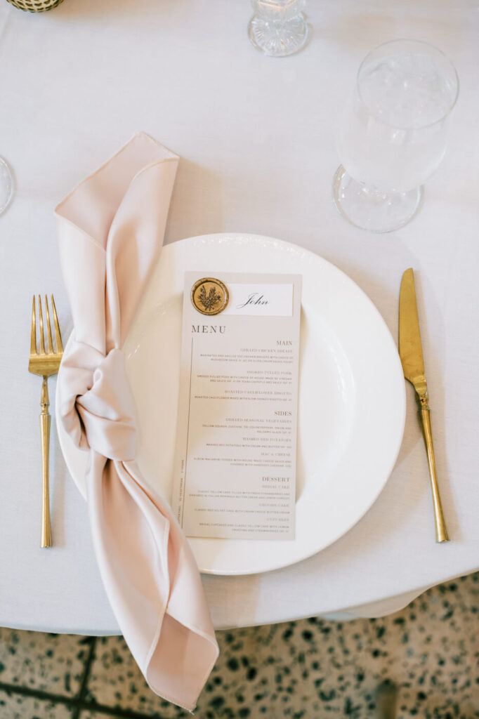 Close-up of a wedding reception place setting with a silky blush napkin and gold details