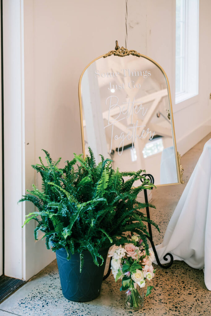 A golden-framed mirror sign with handwritten calligraphy reading "Some things just go better together" sits next to a lush potted fern and floral arrangement.