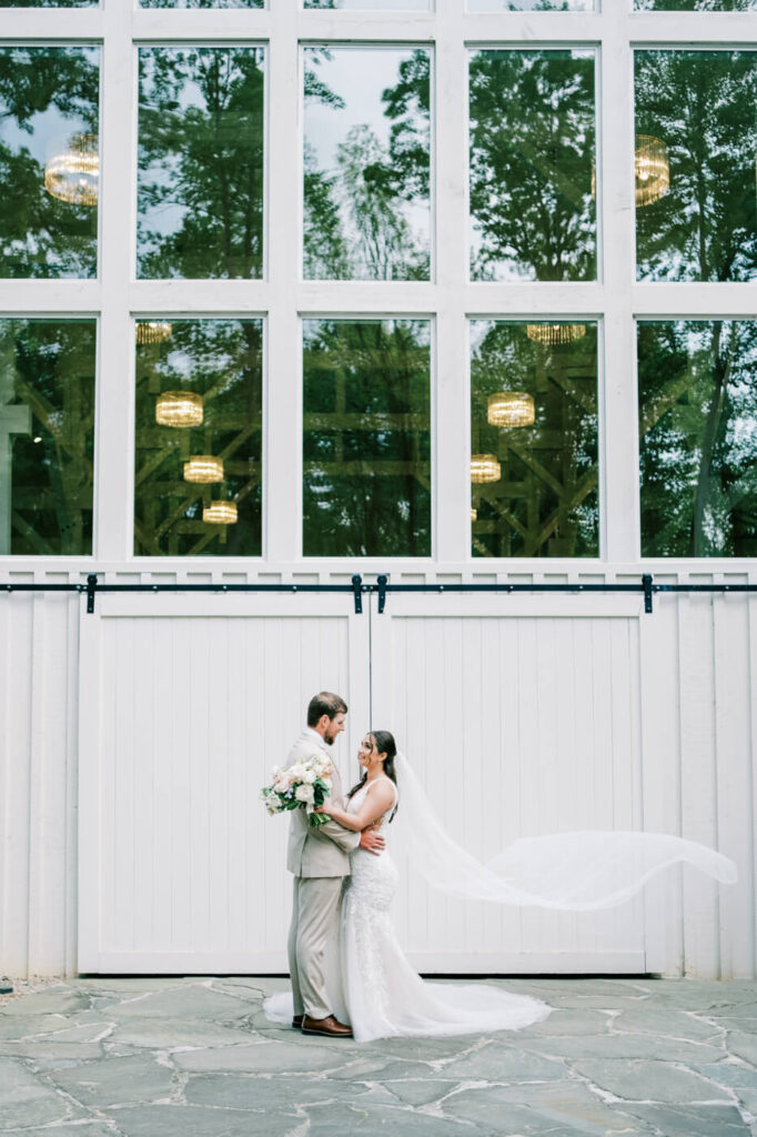 The bride and groom stand close together, gazing into each other's eyes with soft smiles, framed by the elegant white barn backdrop of Carolina Grove.